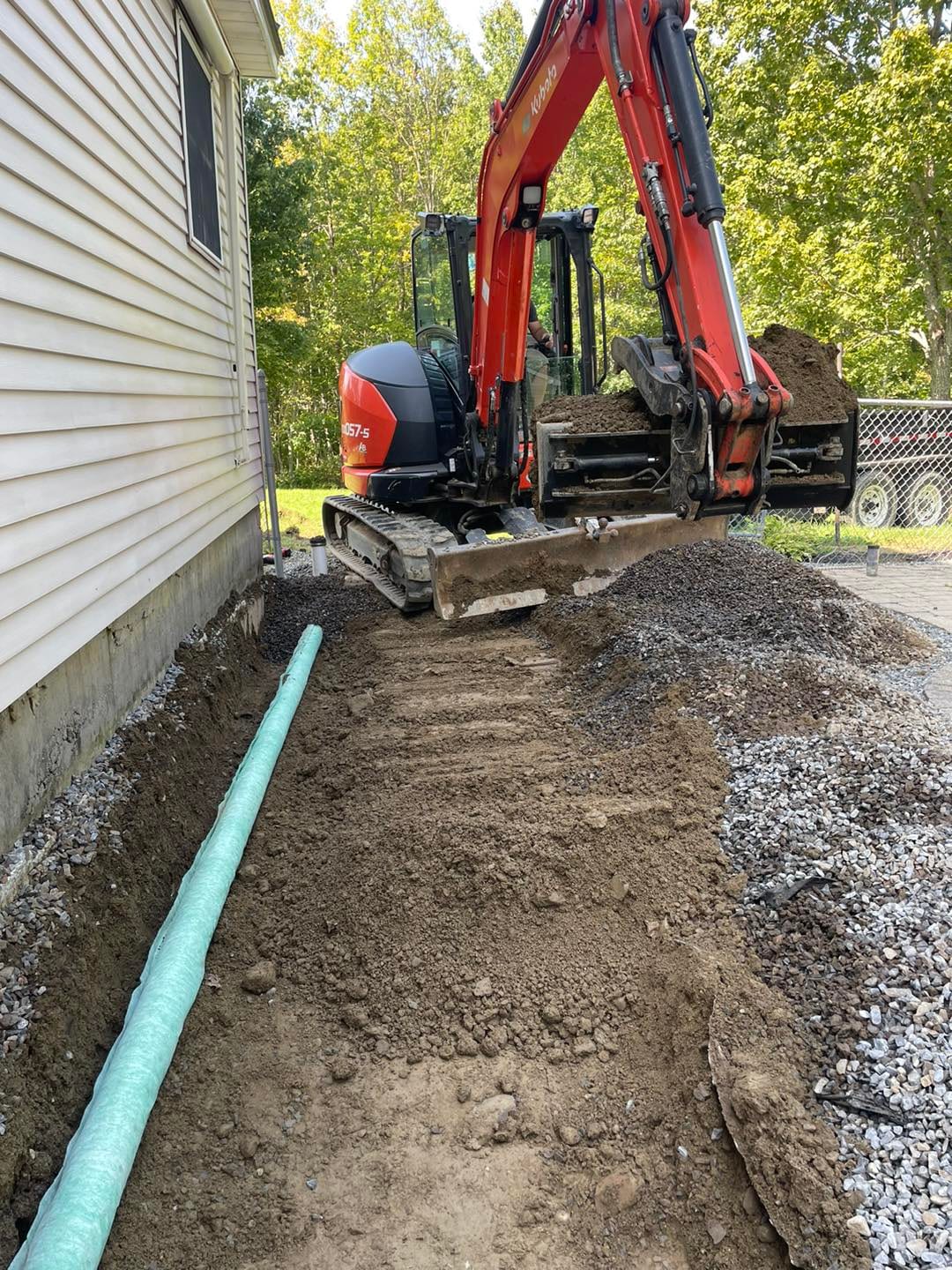 Mini excavator trenching a drain line along the side of a house