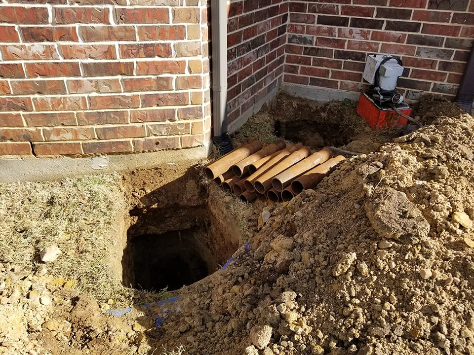 Excavated foundation footing beside a brick home with a hydraulic pump in place